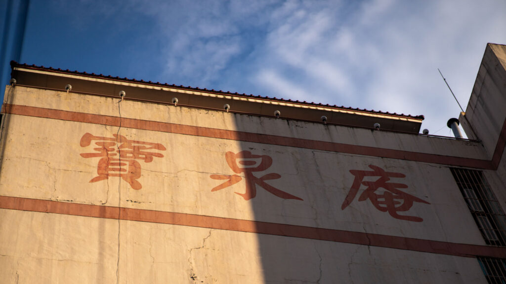 A weathered building wall with large red Chinese characters painted across it, partially covered by a shadow, under a blue sky with scattered clouds.