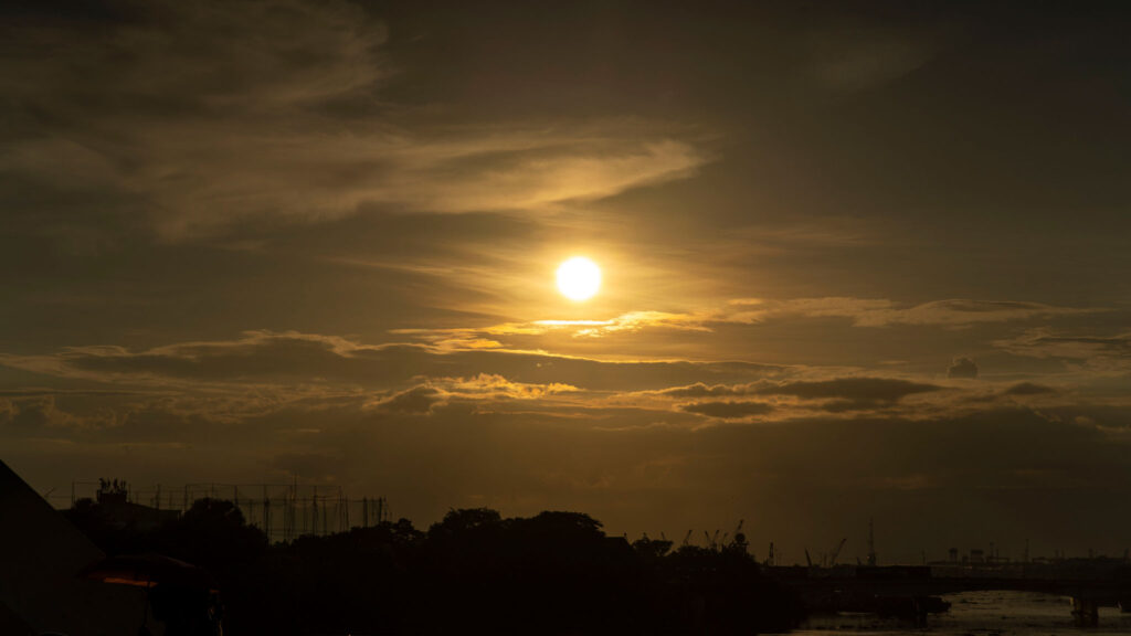Golden sunset over a city skyline with clouds, silhouettes of trees, and distant structures.